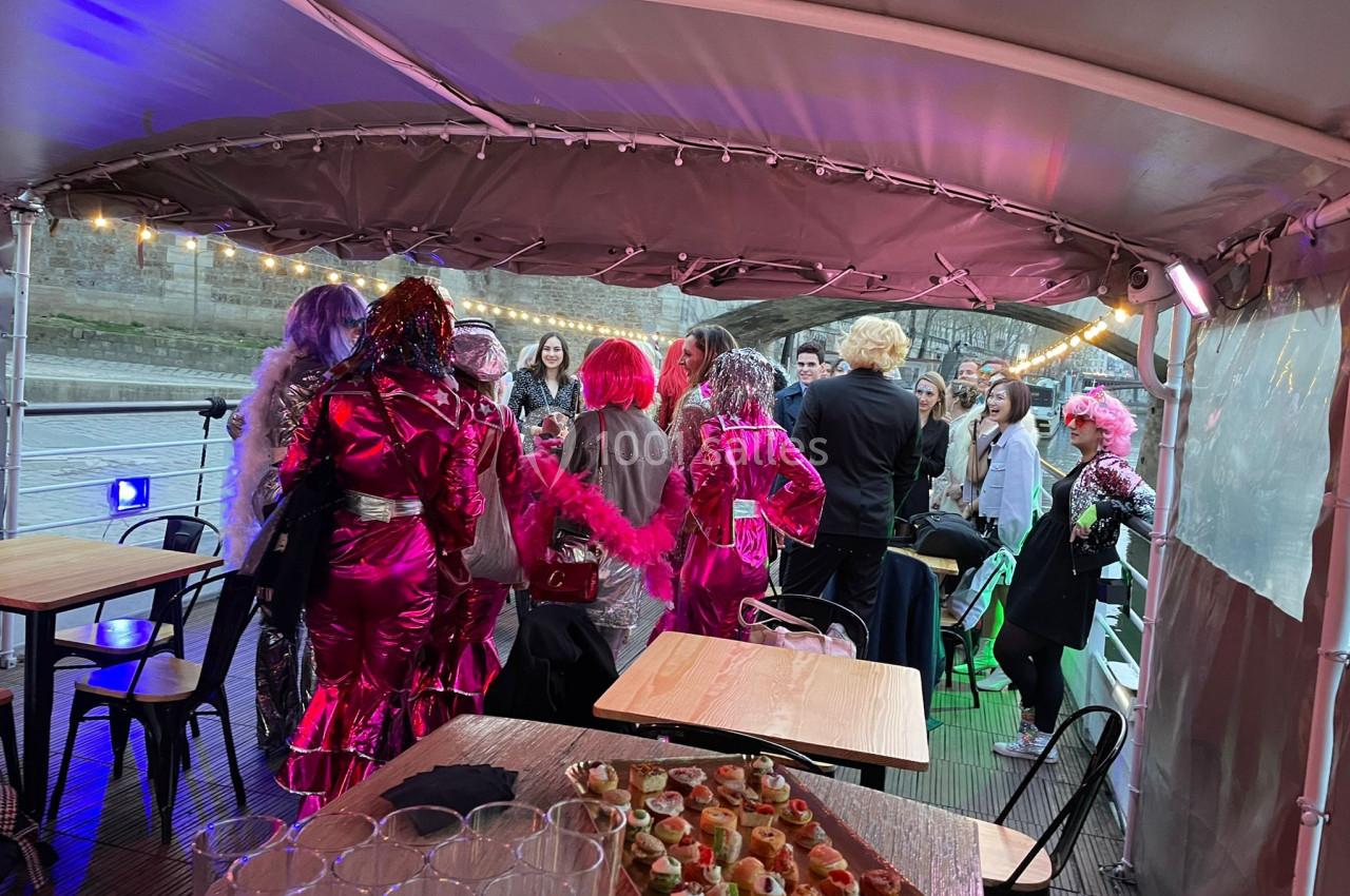 Groupe de personnes en costumes colorés discutant sur une péniche, avec des amuse-bouches et des verres au premier plan.