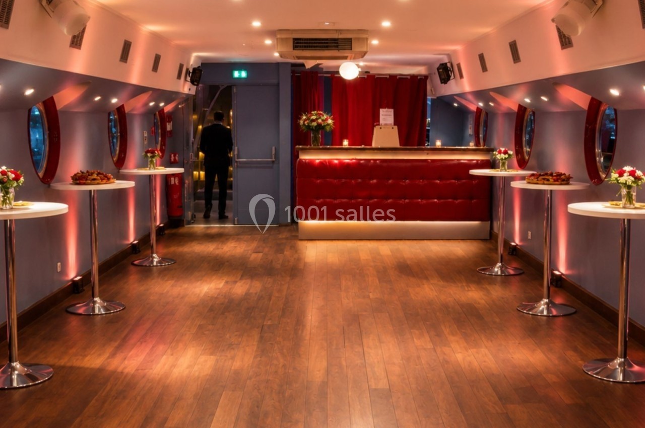 Salle élégante avec parquet, tables hautes décorées de fleurs et un bar rouge au fond, éclairée par une lumière tamisée.