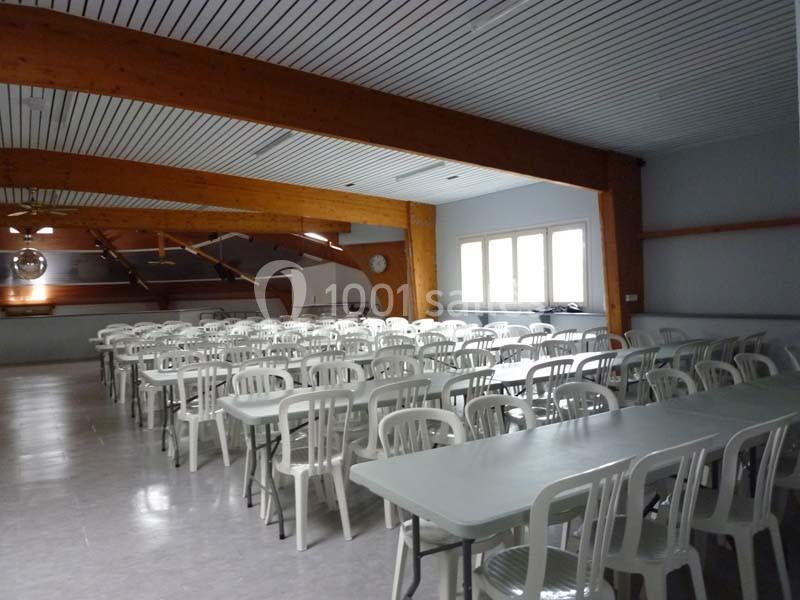 Salle spacieuse avec des tables et des chaises blanches alignées, éclairée par des fenêtres et des luminaires.