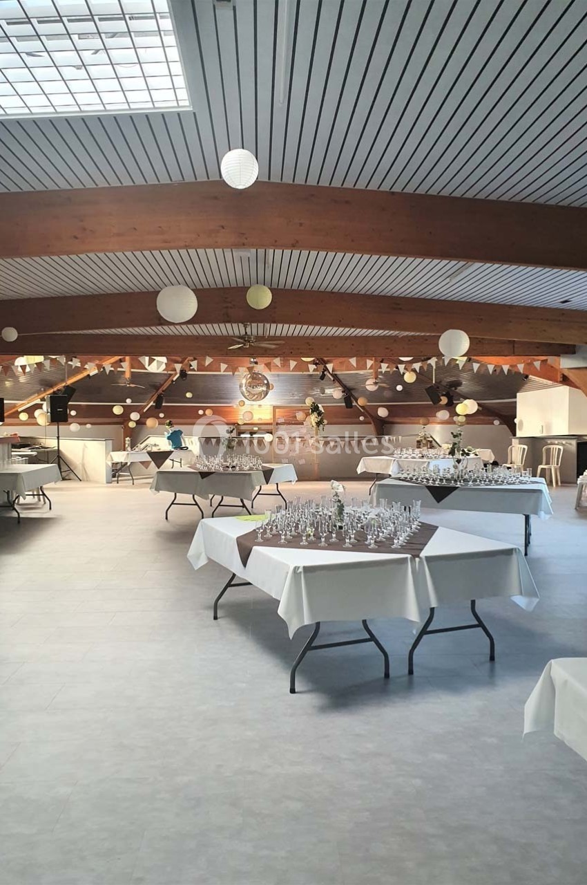 Salle de réception lumineuse avec tables dressées, nappes blanches et décorations suspendues au plafond.