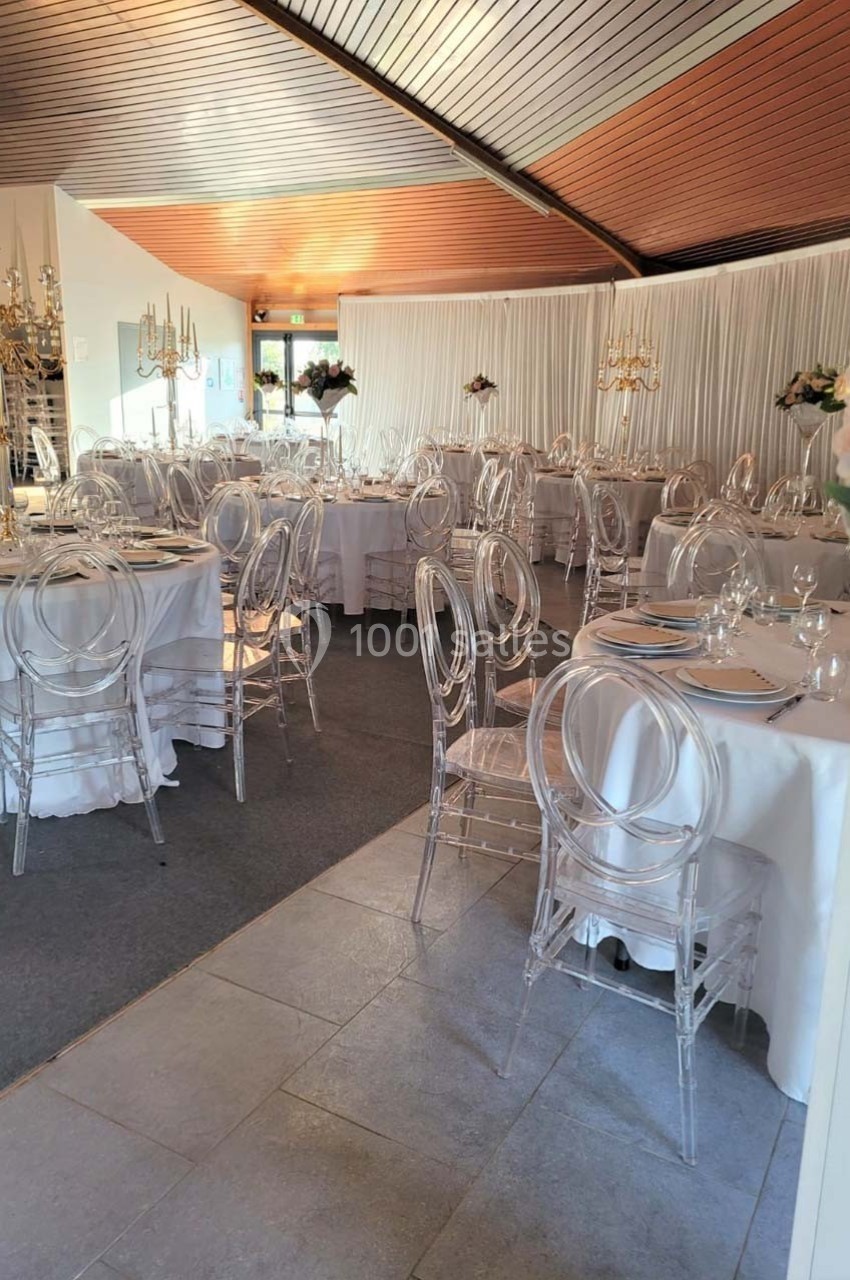 Salle de réception décorée avec des tables rondes, nappes blanches, chaises transparentes et chandeliers dorés.