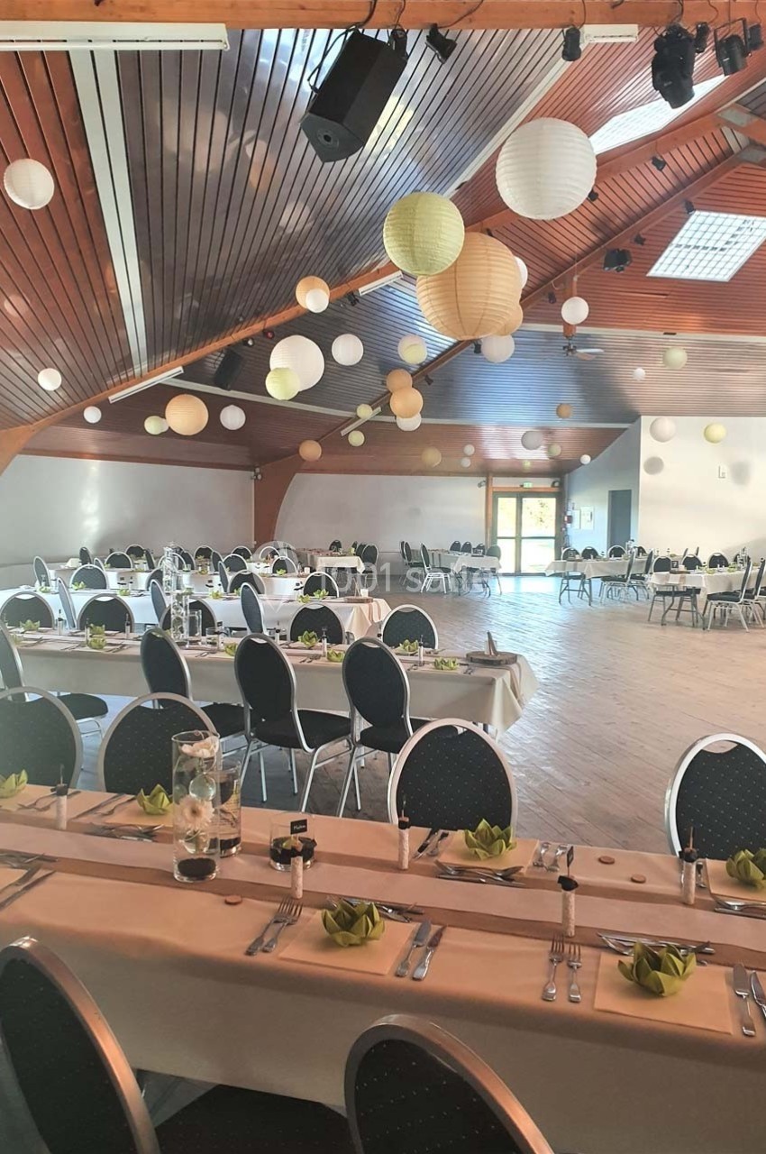Salle de réception décorée avec des lanternes suspendues, tables dressées avec nappes blanches et serviettes vertes.
