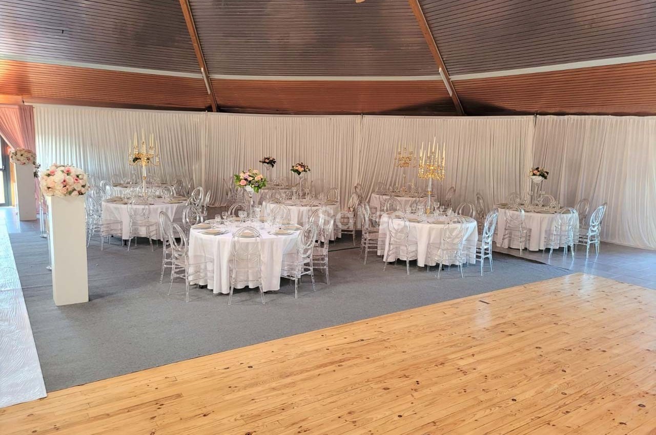 Salle de réception décorée avec des tables rondes, nappes blanches, chaises transparentes et centres de table fleuris.