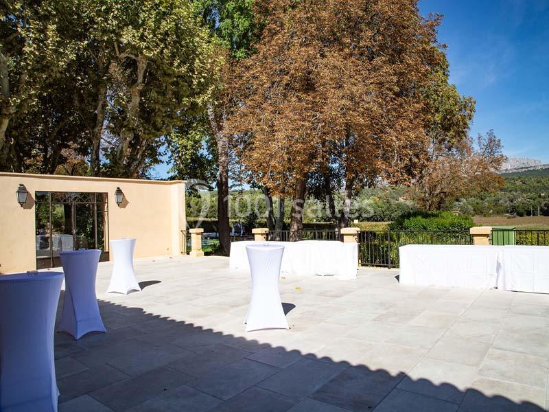 Terrasse ensoleillée avec tables hautes drapées de blanc, entourée d'arbres et d'un paysage verdoyant.