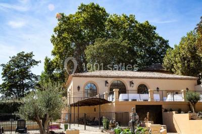 Façade d'une maison méditerranéenne avec terrasse, jardin aménagé et statues, entourée d'arbres sous un ciel dégagé.