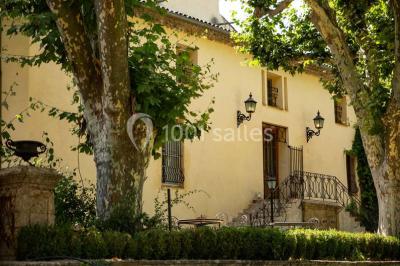 Façade d'une maison méditerranéenne avec terrasse, jardin aménagé et statues, entourée d'arbres sous un ciel dégagé.
