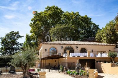Façade d'une maison méditerranéenne avec terrasse, jardin aménagé et statues, entourée d'arbres sous un ciel dégagé.