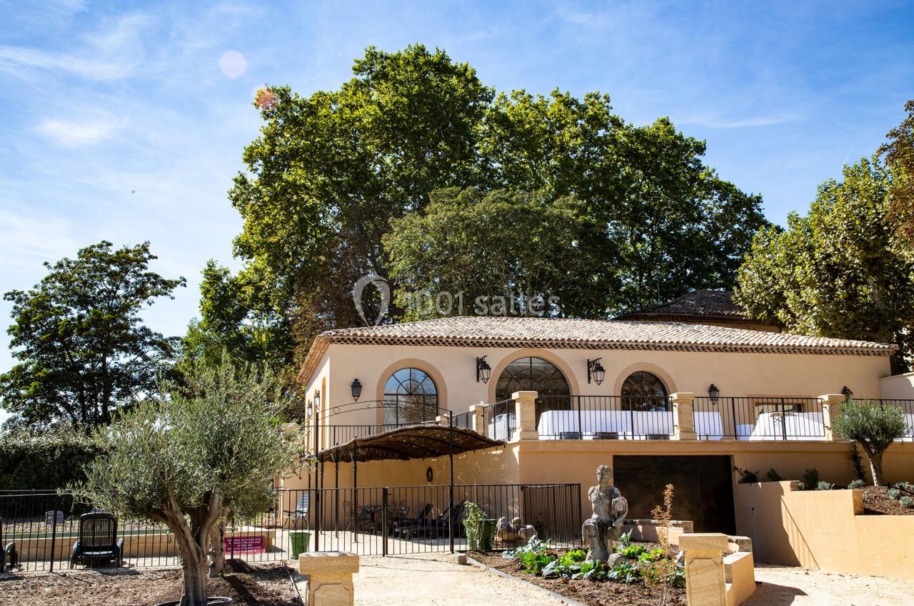 Façade d'une maison méditerranéenne avec terrasse, jardin aménagé et statues, entourée d'arbres sous un ciel dégagé.