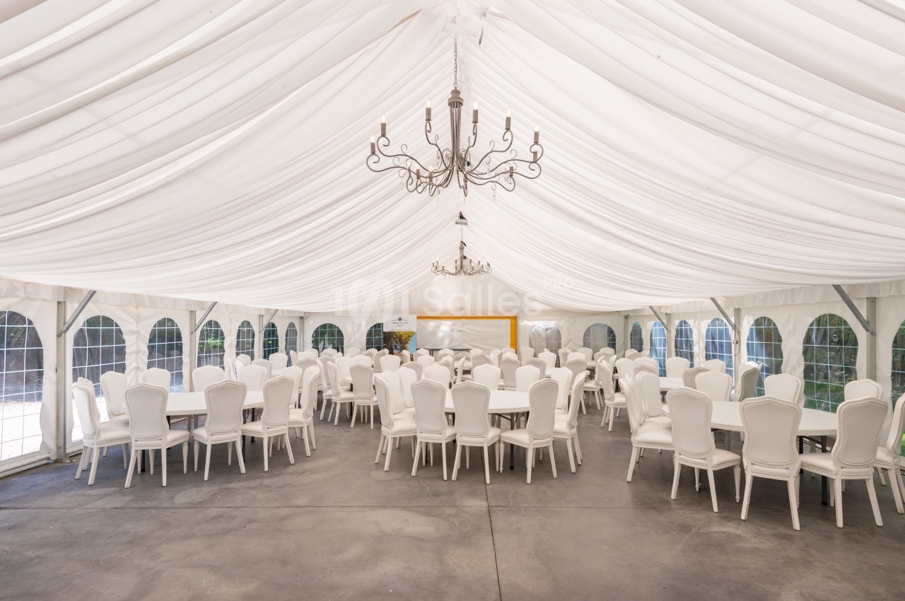 Salle de réception sous une tente blanche avec des tables rondes et des chaises blanches disposées en rangées.