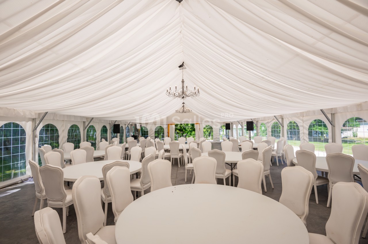 Salle de réception sous une tente blanche avec tables rondes, chaises blanches et lustre central.