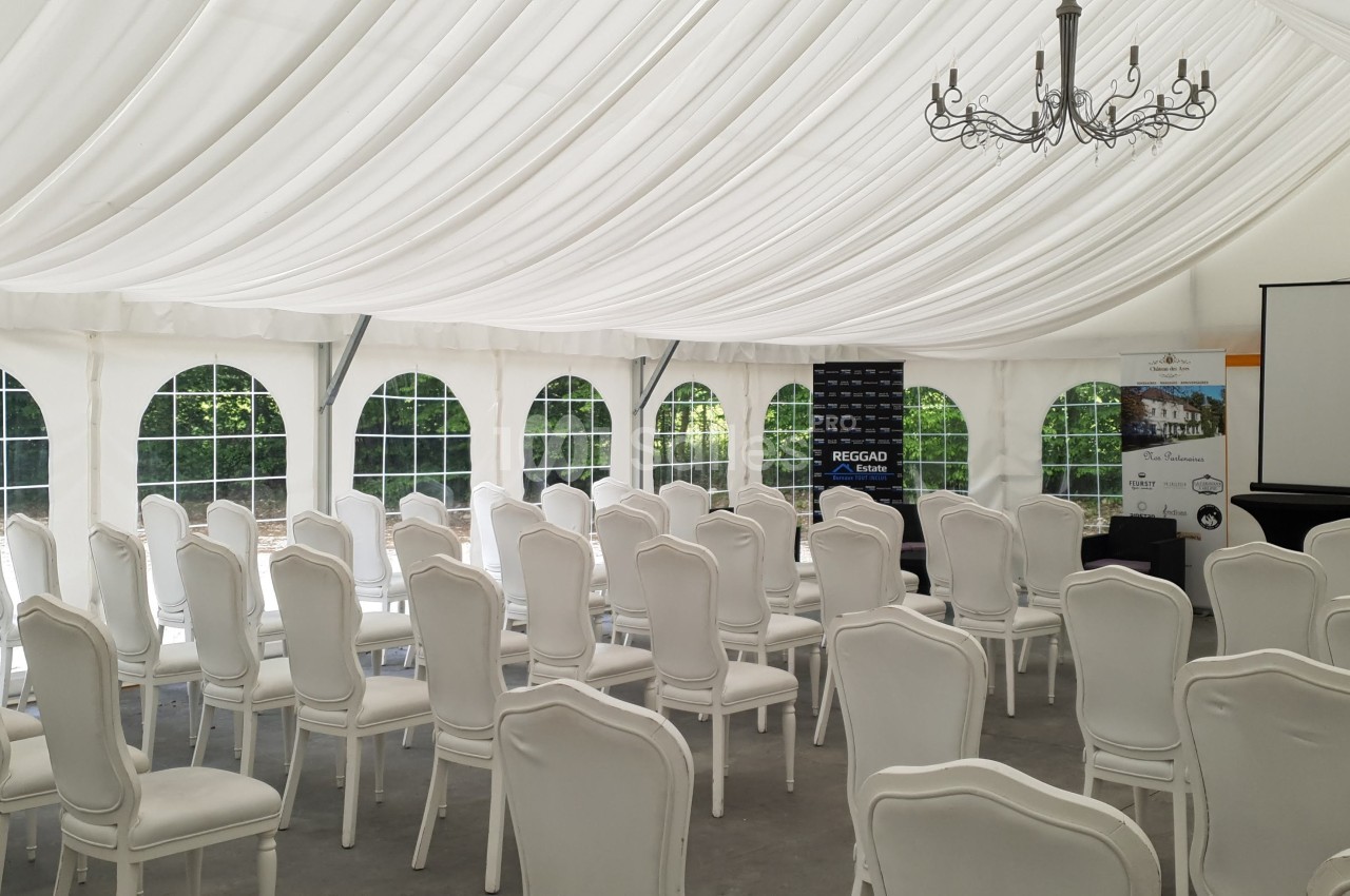 Salle de conférence sous une tente blanche avec des chaises alignées, un écran de projection et un chandelier suspendu.