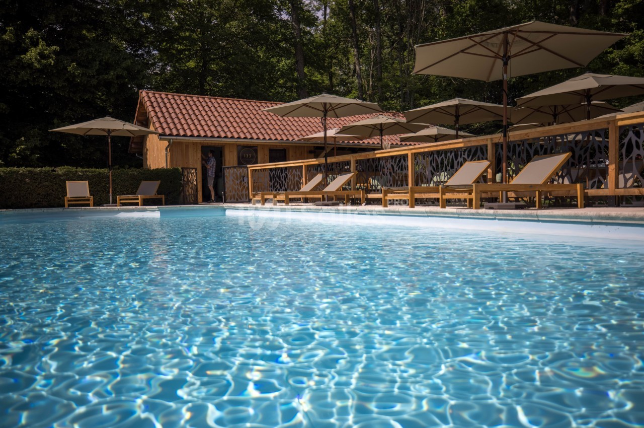 Piscine extérieure entourée de transats en bois et de parasols, avec un bâtiment en bois en arrière-plan.