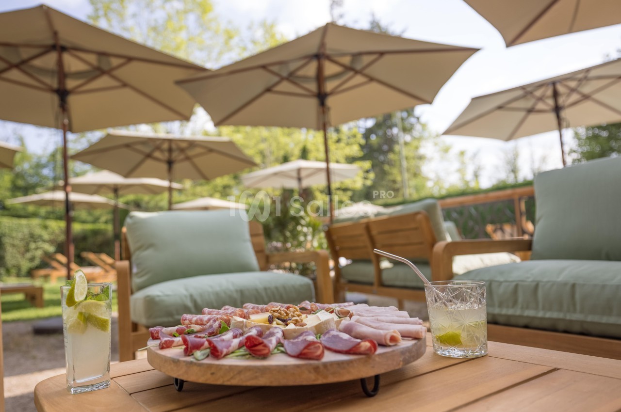 Plateau de charcuterie et fromages sur une table en bois, entouré de fauteuils et parasols dans un jardin ensoleillé.