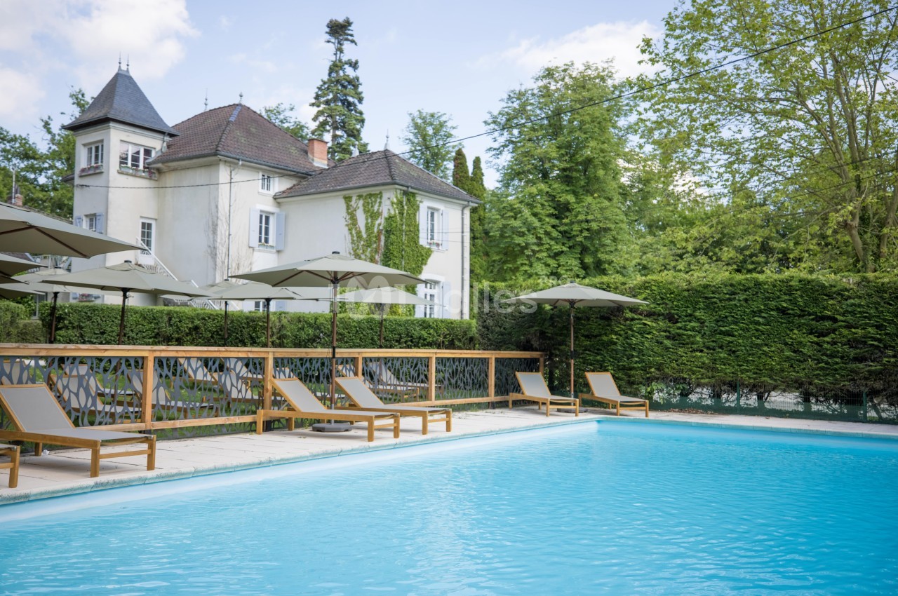 Piscine extérieure entourée de chaises longues et parasols, avec une maison ancienne et des arbres en arrière-plan.
