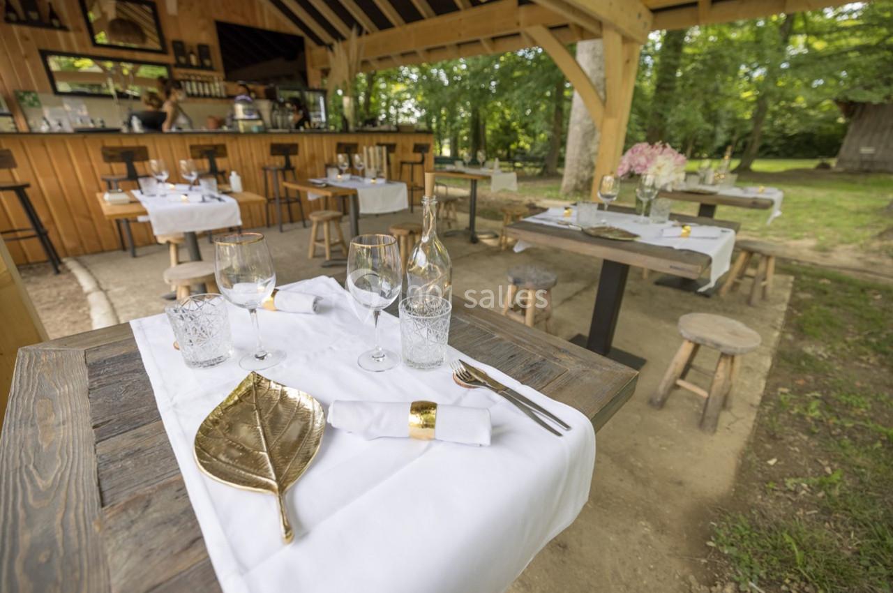 Tables dressées avec nappes blanches, couverts dorés et verres, dans un restaurant en plein air entouré de verdure.