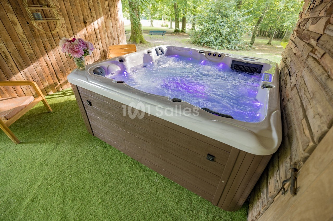 Spa extérieur à remous sur une terrasse en gazon artificiel, entouré de bois et de verdure.