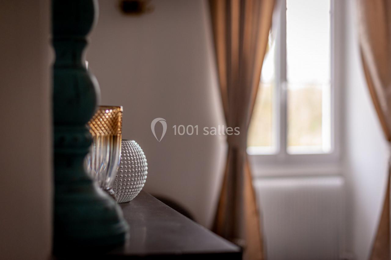 Vase texturé et verre doré posés sur un meuble sombre, devant une fenêtre avec rideaux beiges.