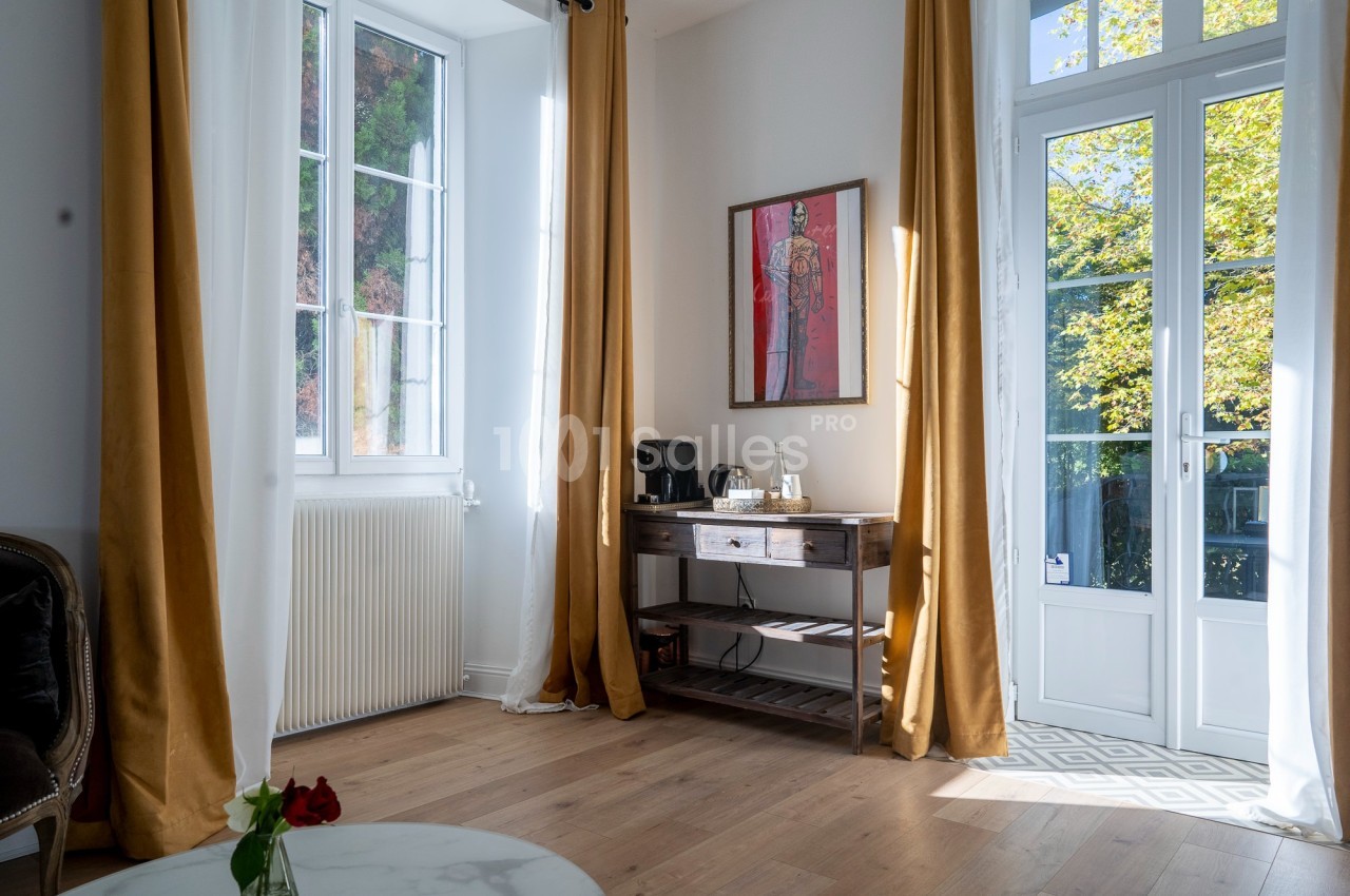 Salon lumineux avec parquet, rideaux dorés, table en bois avec machine à café, et porte-fenêtre donnant sur un jardin.