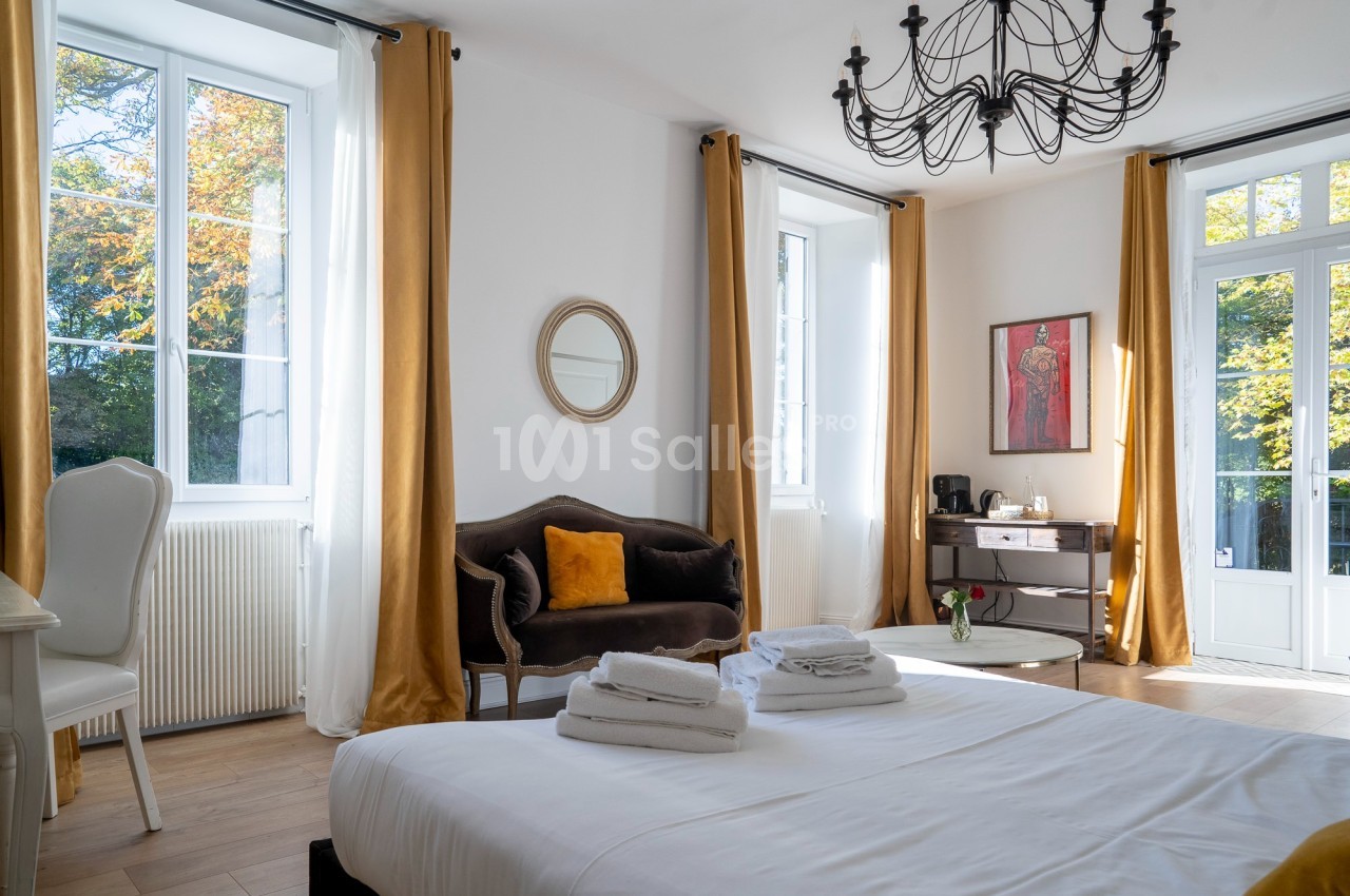 Chambre lumineuse avec lit double, canapé, bureau, grandes fenêtres, rideaux dorés et lustre noir.