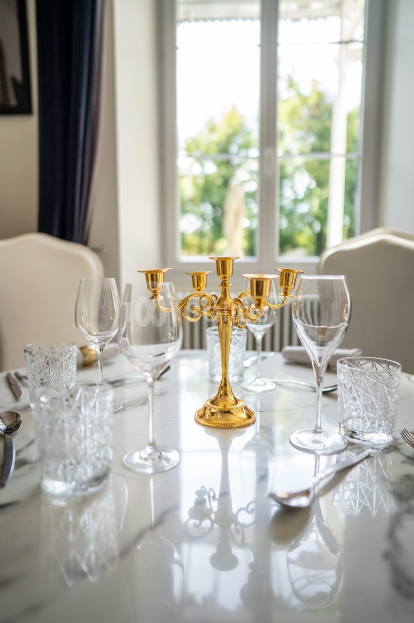 Table dressée avec un chandelier doré, verres et couverts sur une nappe blanche, devant une fenêtre lumineuse.