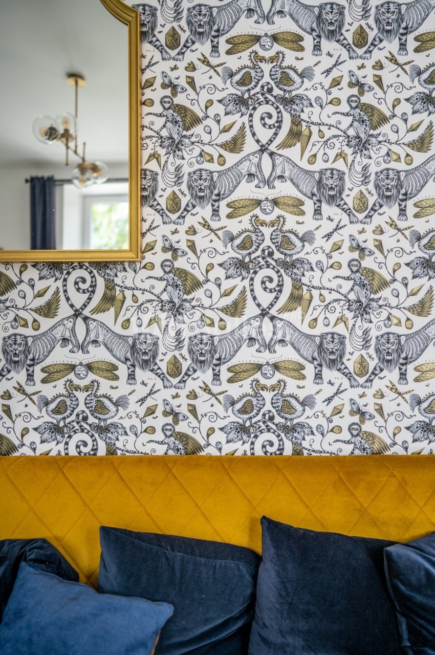 Papier peint à motifs animaliers et végétaux en noir, blanc et doré, avec un canapé jaune et des coussins bleus au premier…
