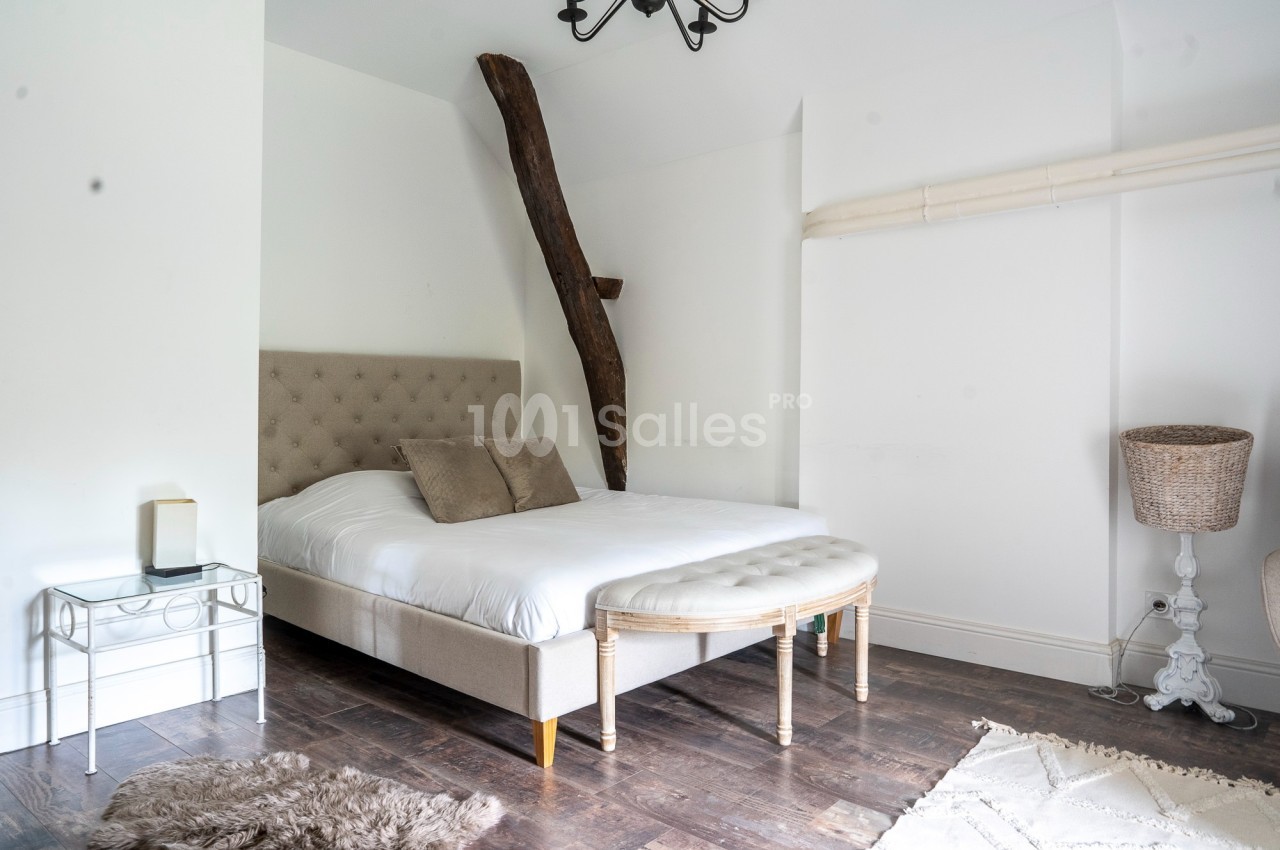 Chambre lumineuse avec un lit double, poutre apparente, sol en bois foncé et mobilier simple.
