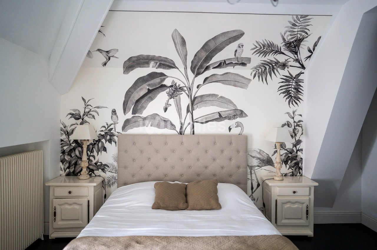 Chambre lumineuse avec un lit double, tête de lit capitonnée, tables de chevet et mur décoré d'une fresque végétale.