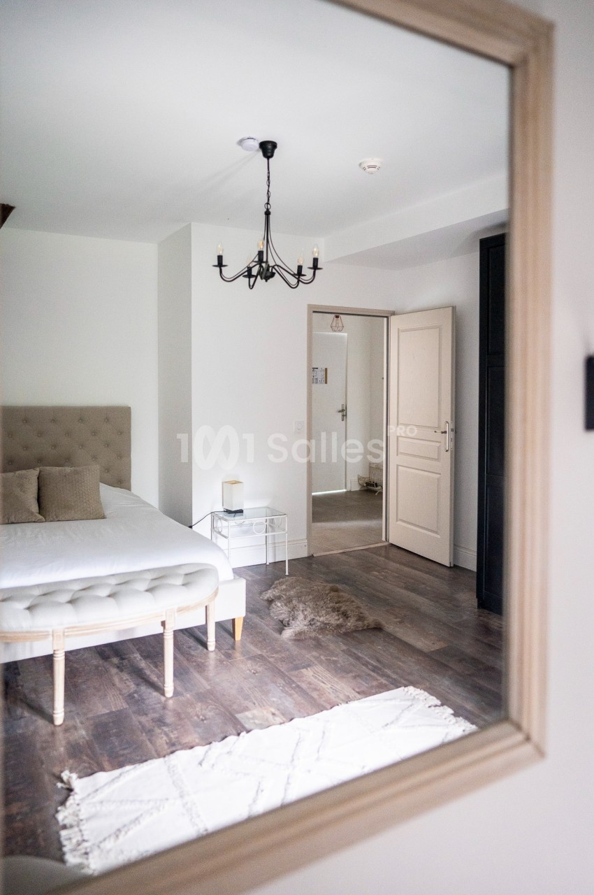 Chambre lumineuse avec lit double, sol en bois, tapis clair, miroir et porte ouverte sur un couloir.