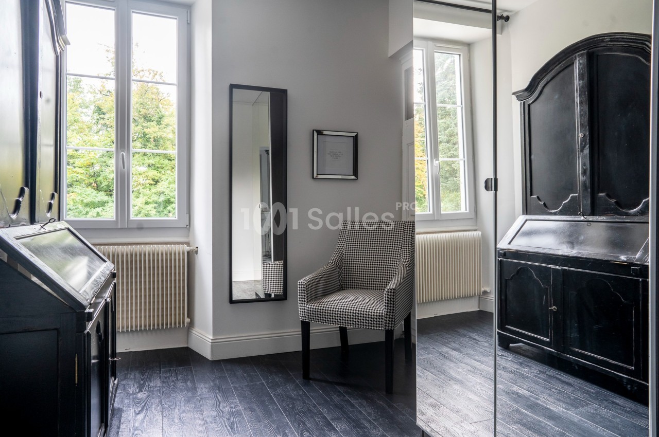 Pièce lumineuse avec un fauteuil à motif, un miroir, un meuble noir et des fenêtres donnant sur un espace vert.