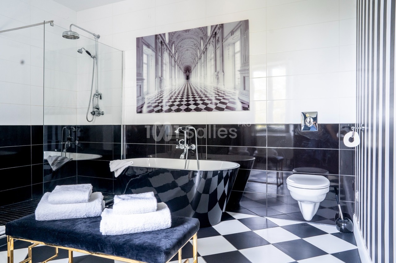 Salle de bain moderne avec baignoire noire, douche vitrée, toilettes suspendues et sol en damier noir et blanc.
