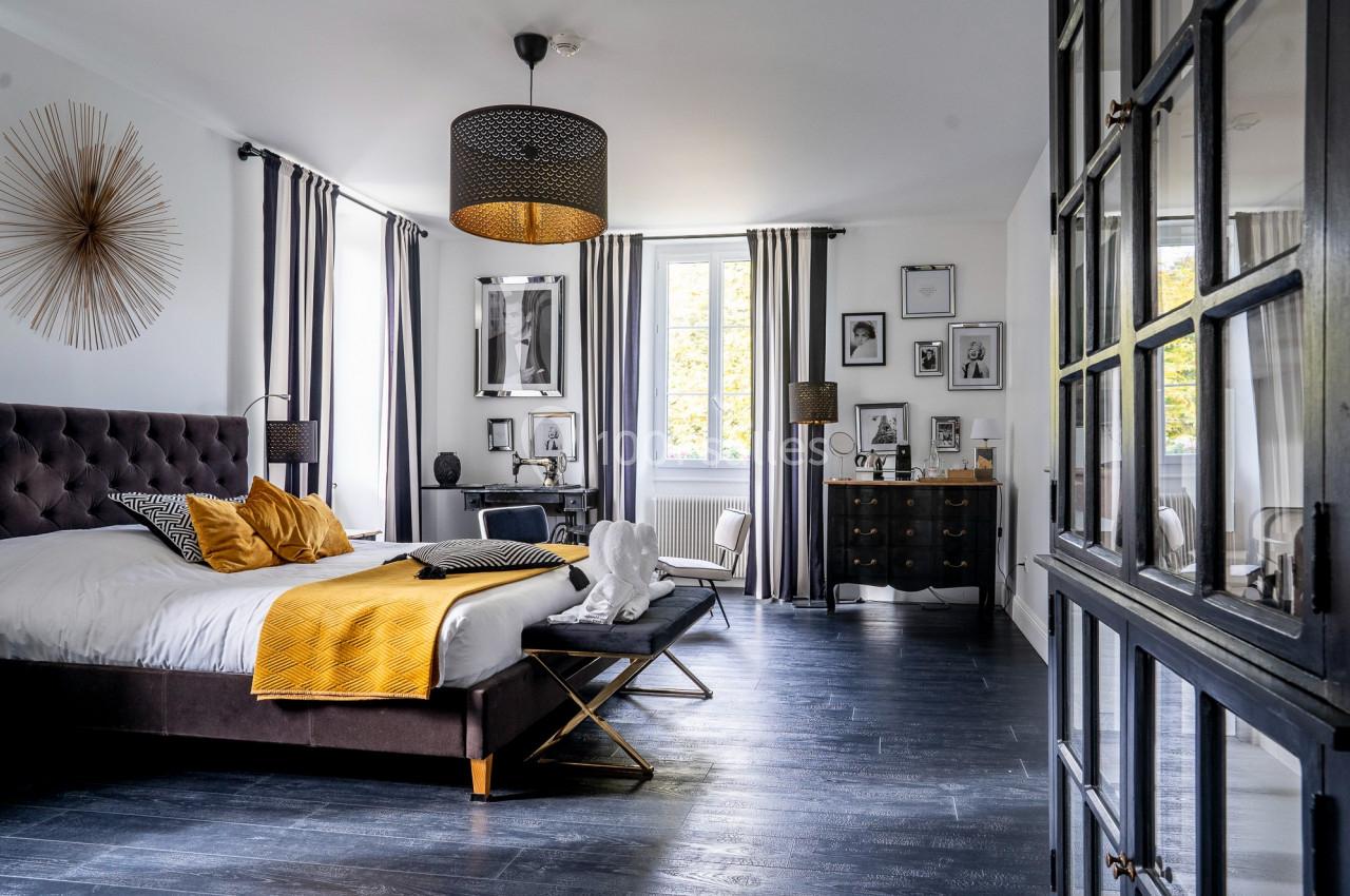 Chambre lumineuse avec lit double, décoration moderne, parquet sombre, murs blancs et grande fenêtre avec rideaux.