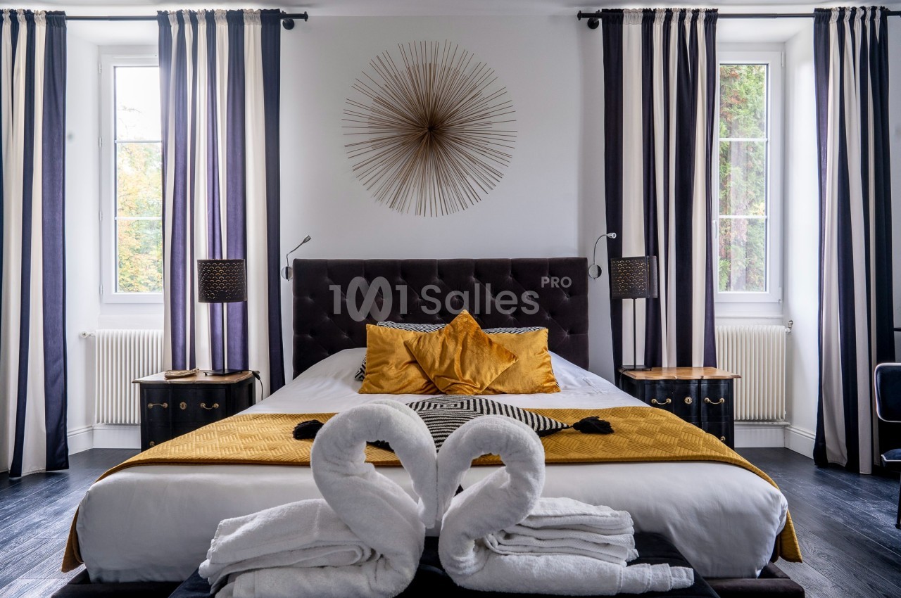 Chambre élégante avec lit double, coussins dorés, serviettes pliées en cygnes et grandes fenêtres encadrées de rideaux rayés.