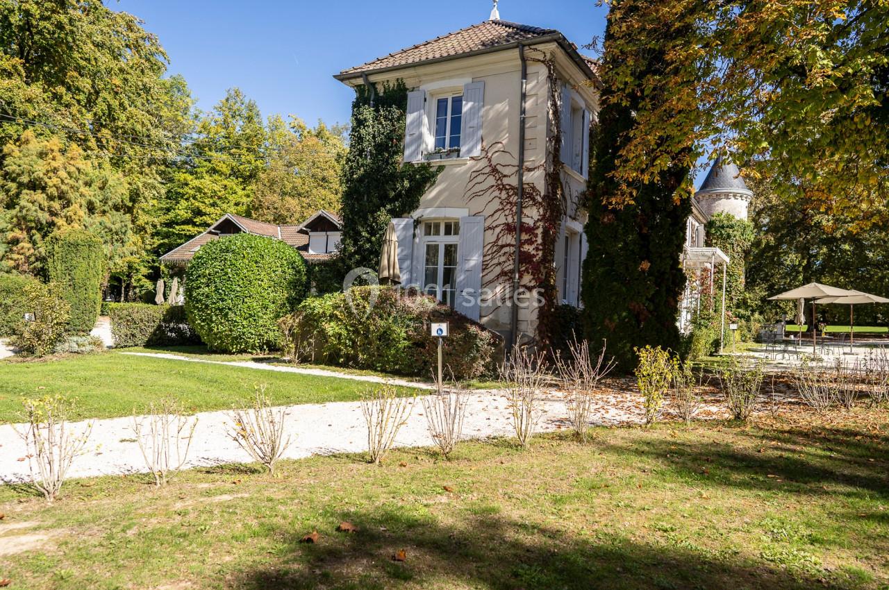 Maison entourée de verdure avec un chemin en gravier, des arbres et une terrasse visible en arrière-plan.