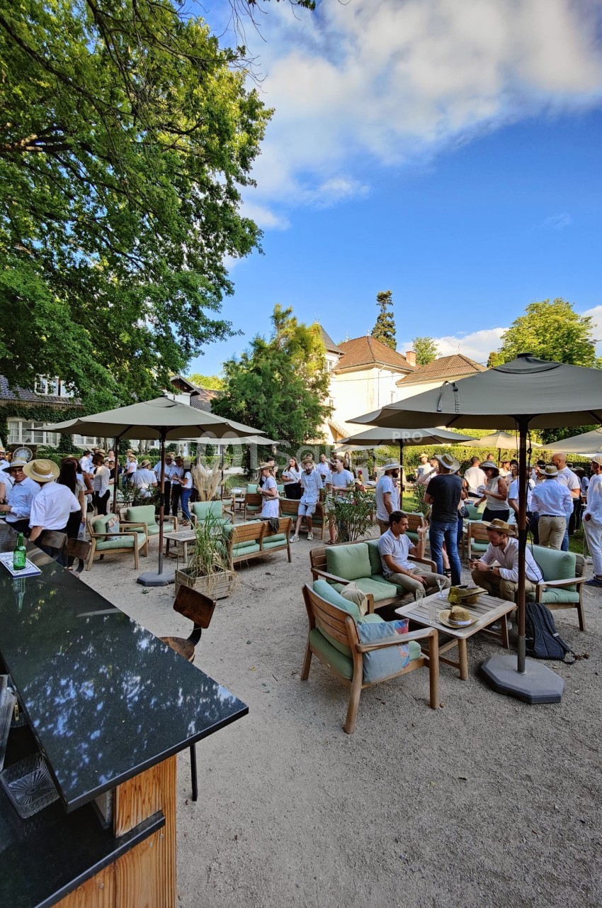 Groupe de personnes rassemblées dans un jardin avec des parasols, des tables et des chaises, par une journée ensoleillée.