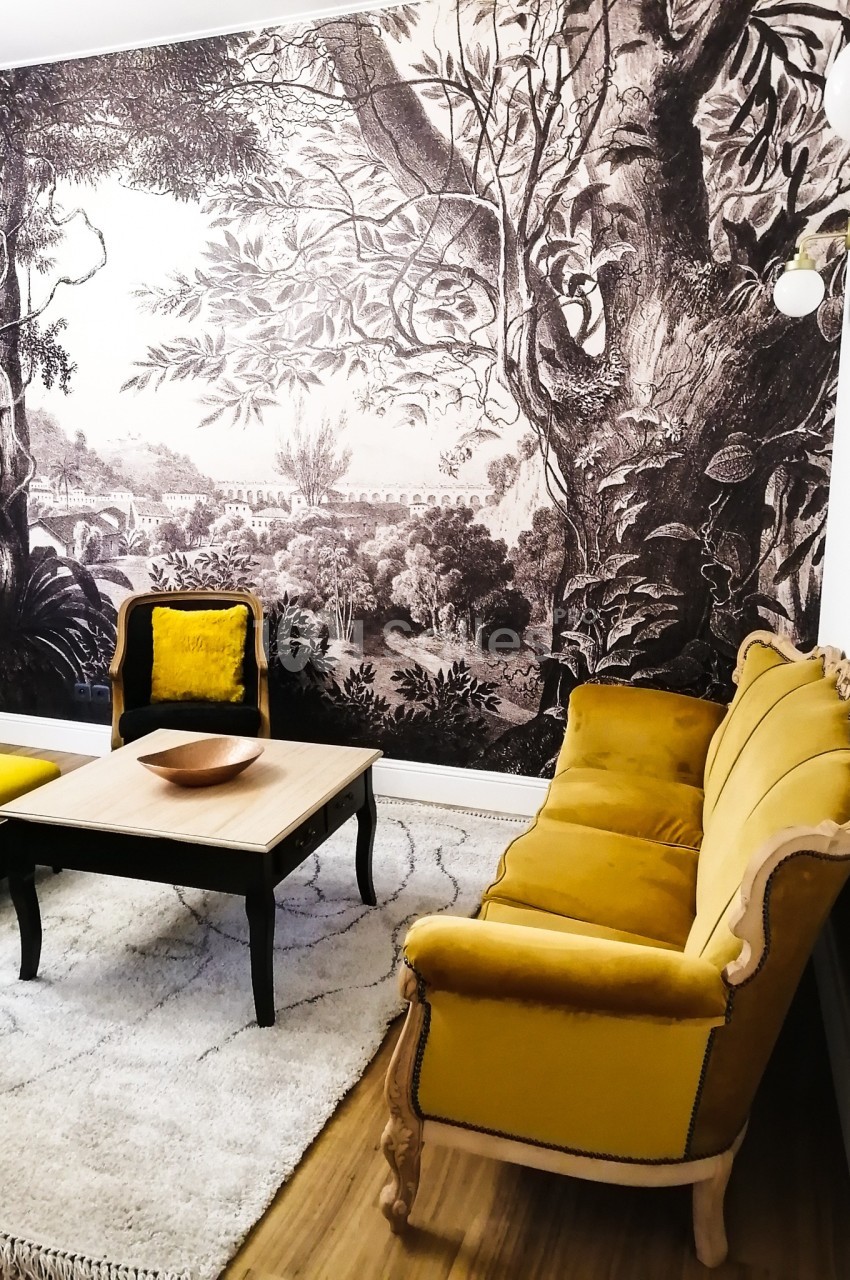 Salon avec canapé et fauteuils jaunes, table basse en bois, tapis clair et mur décoré d'une fresque végétale en noir et…