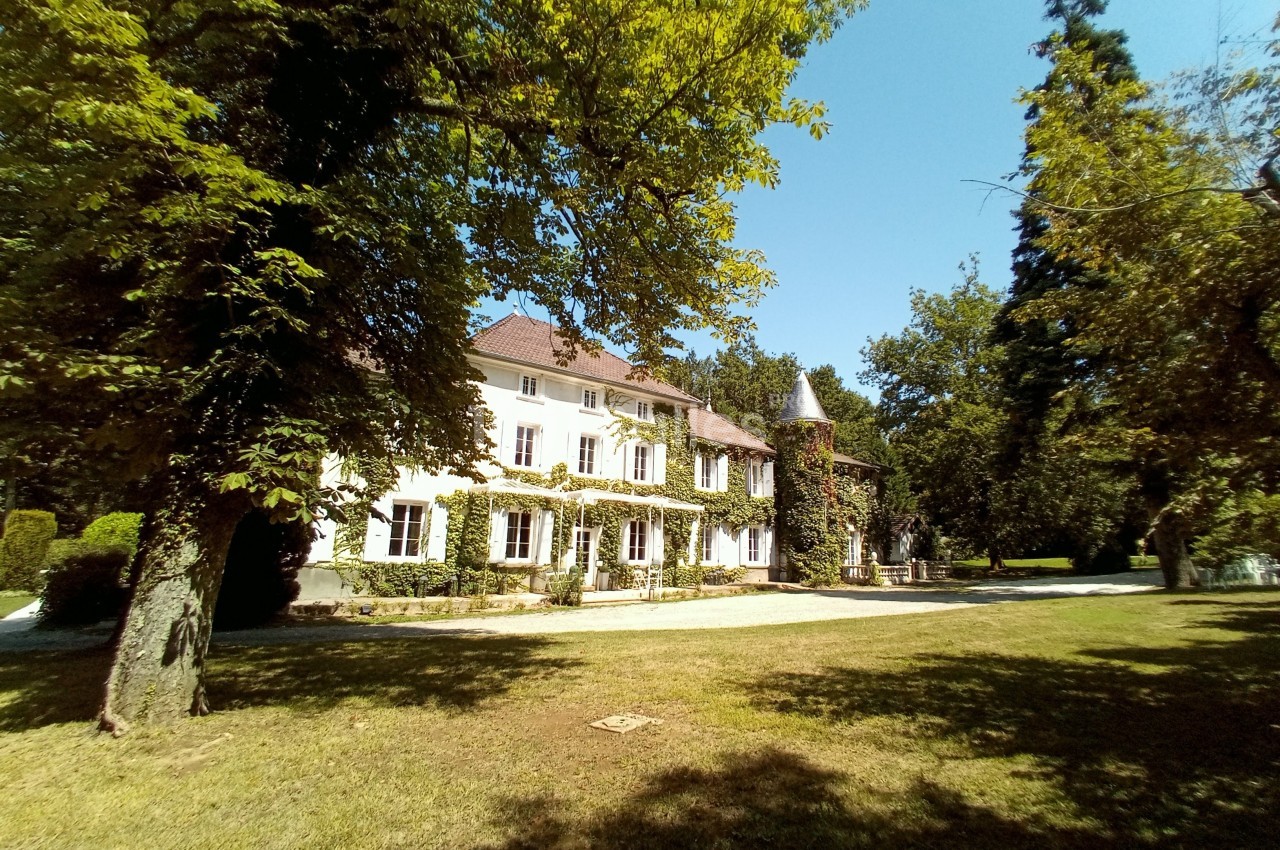 Grande maison blanche entourée d'arbres, avec une tour en pierre et une pelouse au premier plan.