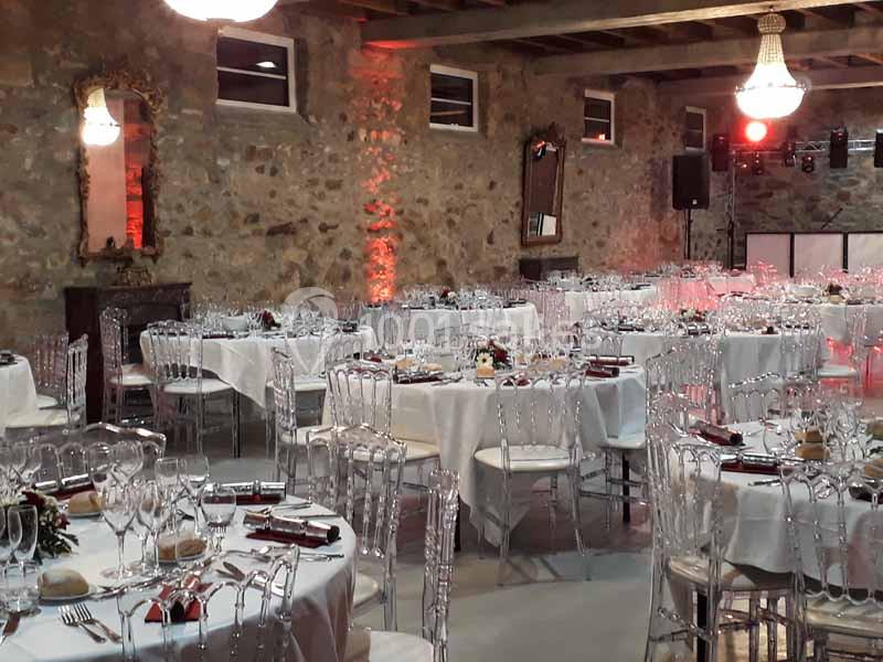 Salle de réception en pierre avec tables dressées, chaises transparentes et éclairage chaleureux.