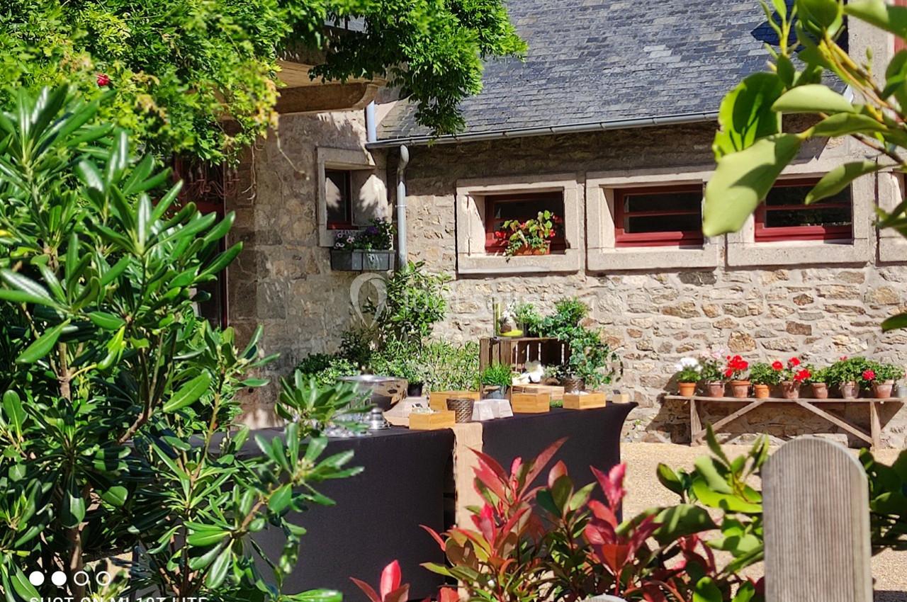 Cour extérieure ensoleillée avec des tables dressées, entourée de plantes, d'arbustes et d'un bâtiment en pierre.