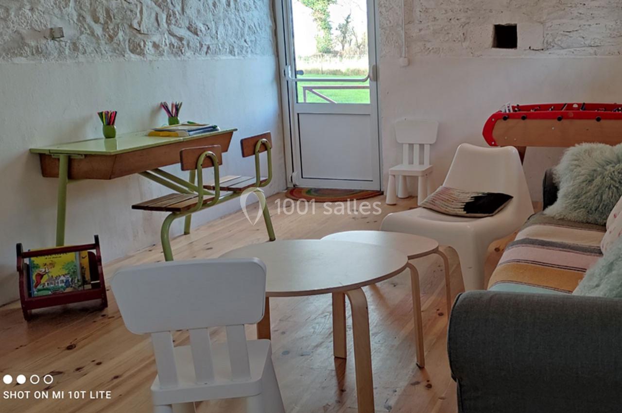 Salle lumineuse avec mobilier simple : table, chaises, canapé, baby-foot et vue sur un jardin par une porte vitrée.