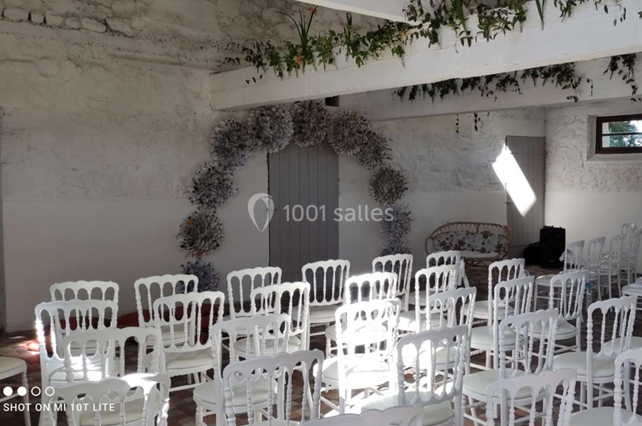 Salle décorée pour un événement, avec des chaises blanches alignées et une arche florale devant une porte grise.