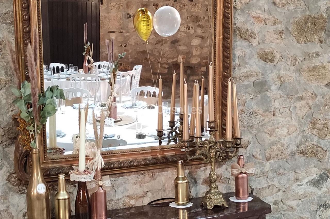 Décoration de table avec bougies dorées, bouteilles décoratives et ballons reflétés dans un miroir sur un mur en pierre.