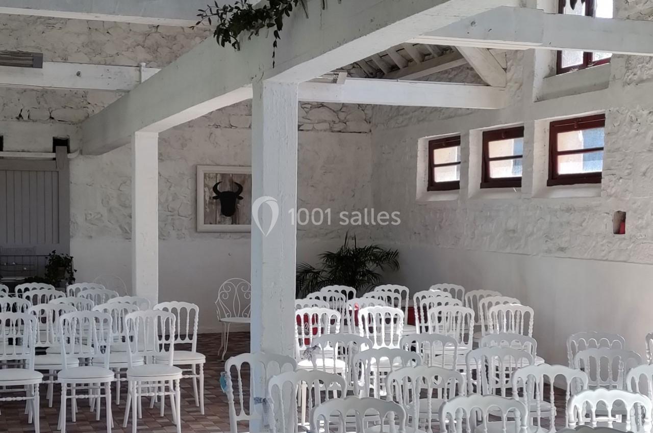 Salle lumineuse avec des murs en pierre blanche, des chaises blanches alignées et des poutres apparentes.