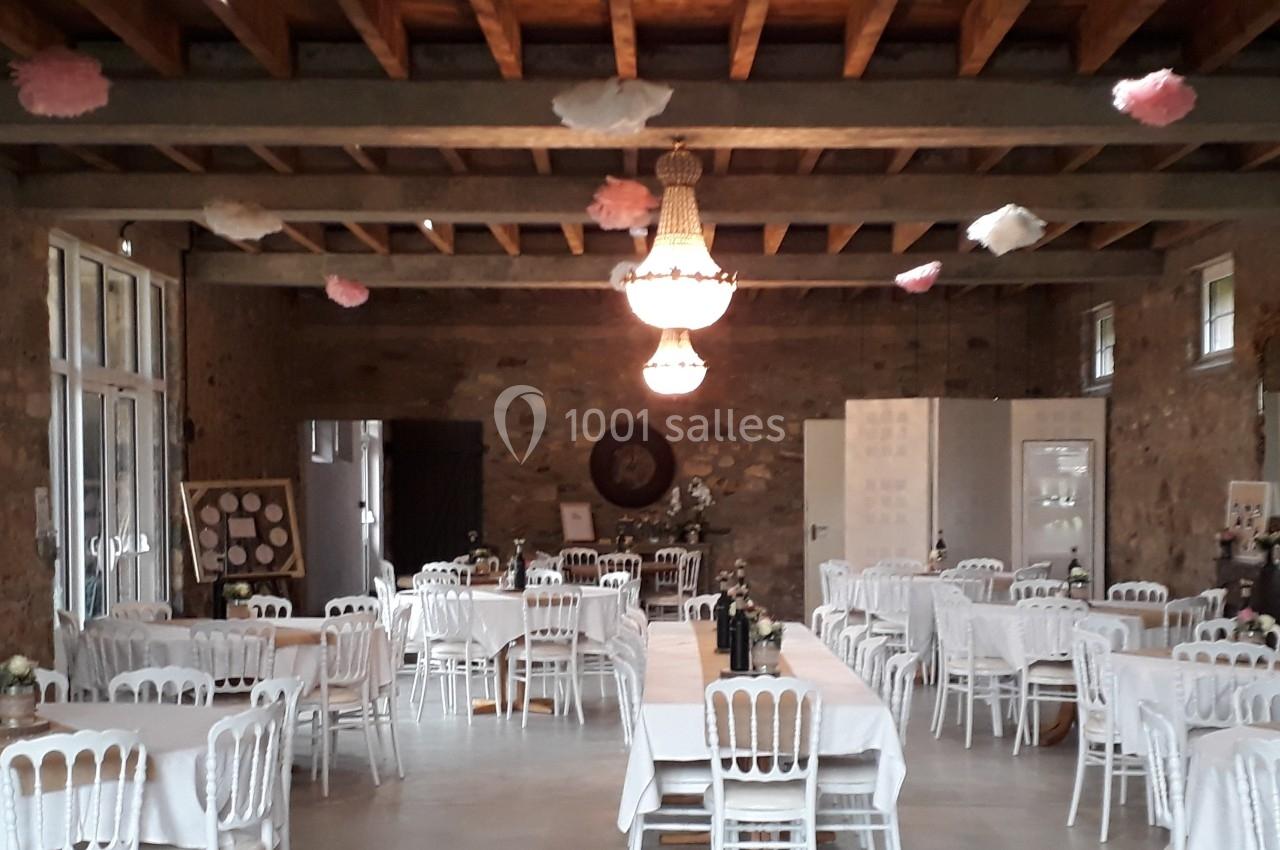 Salle de réception avec tables dressées, chaises blanches et décoration suspendue sous un plafond en poutres apparentes.