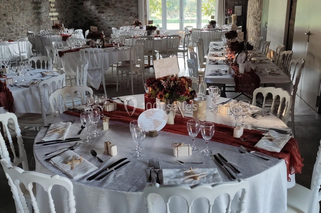 Salle de réception décorée pour un événement, avec tables rondes dressées, nappes blanches et centres de table fleuris.