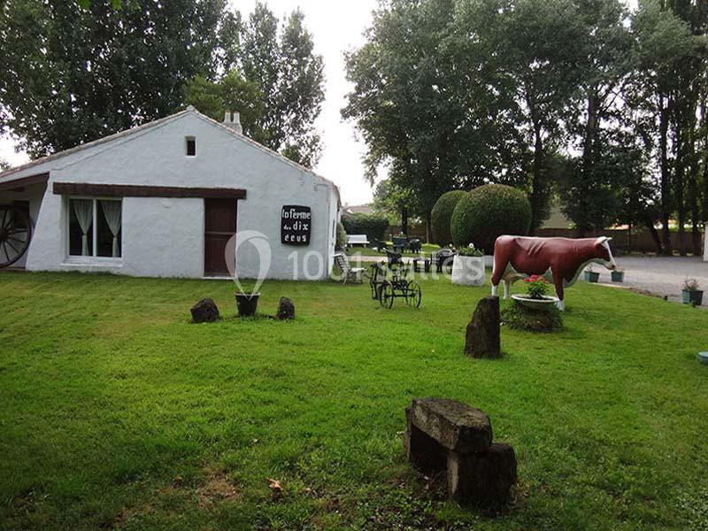 Maison blanche avec jardin verdoyant, tables extérieures, statue de vache rouge et arbres en arrière-plan.