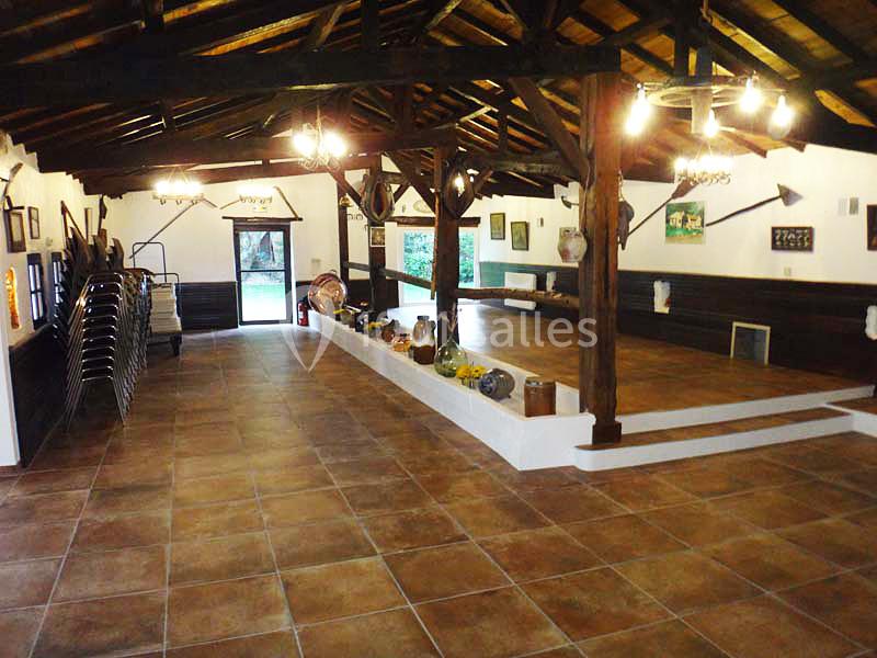 Salle intérieure spacieuse avec plafond en bois, carrelage au sol, objets décoratifs et chaises empilées sur le côté.