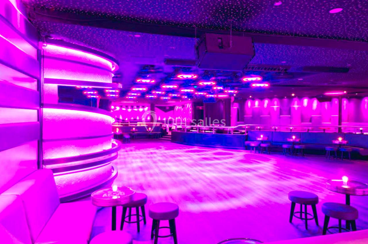 Salle de danse vide éclairée par des lumières violettes, avec des tables et des sièges disposés autour.