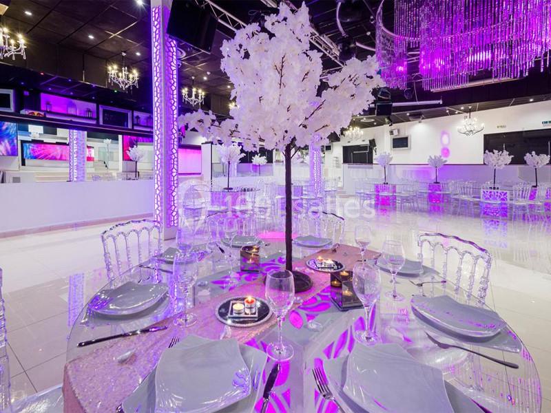 Salle de réception décorée avec des tables rondes, des chaises transparentes et un arbre central blanc sous un éclairage…