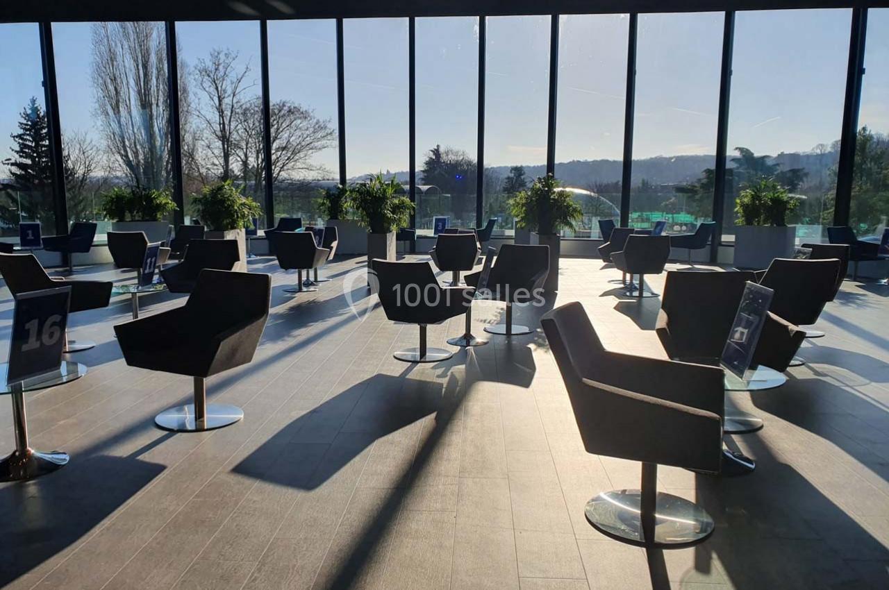 Salle lumineuse avec de grandes baies vitrées, des fauteuils noirs espacés et des plantes disposées près des fenêtres.