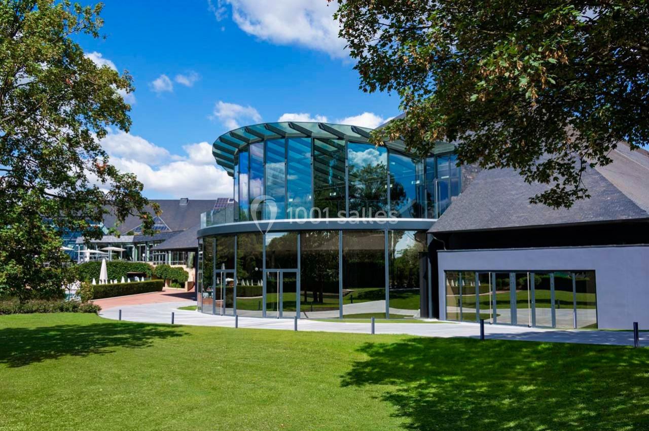 Bâtiment moderne avec façade vitrée arrondie, entouré d'une pelouse et d'arbres sous un ciel bleu.